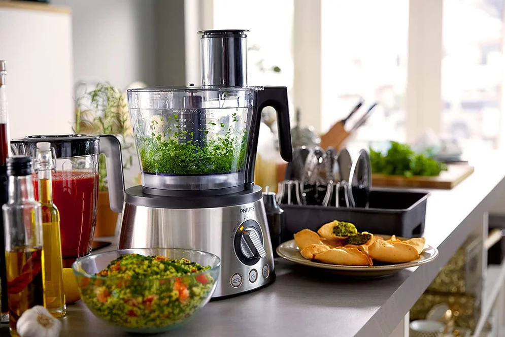 Philips Food Processor – Herb Chopping and Food Preparation Philips food processor with chopped green herbs in its bowl, beside a blender jug and prepared food
